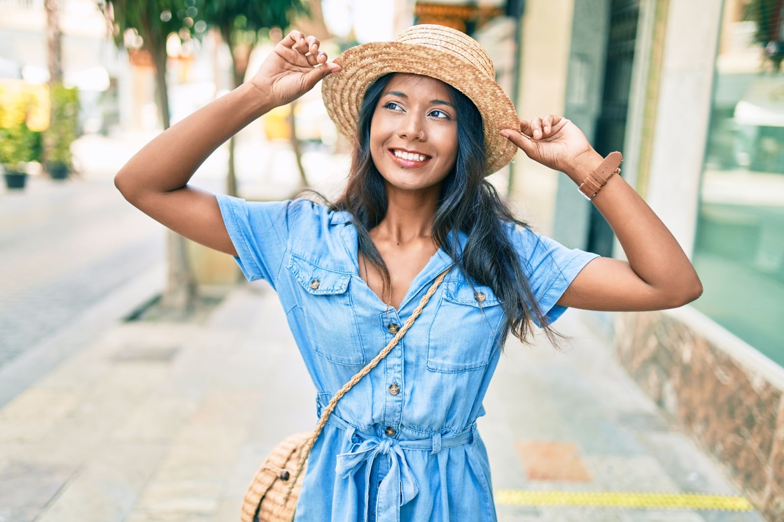 Young woman in a blue shirt dress wearing a summer hat while smiling in the city.